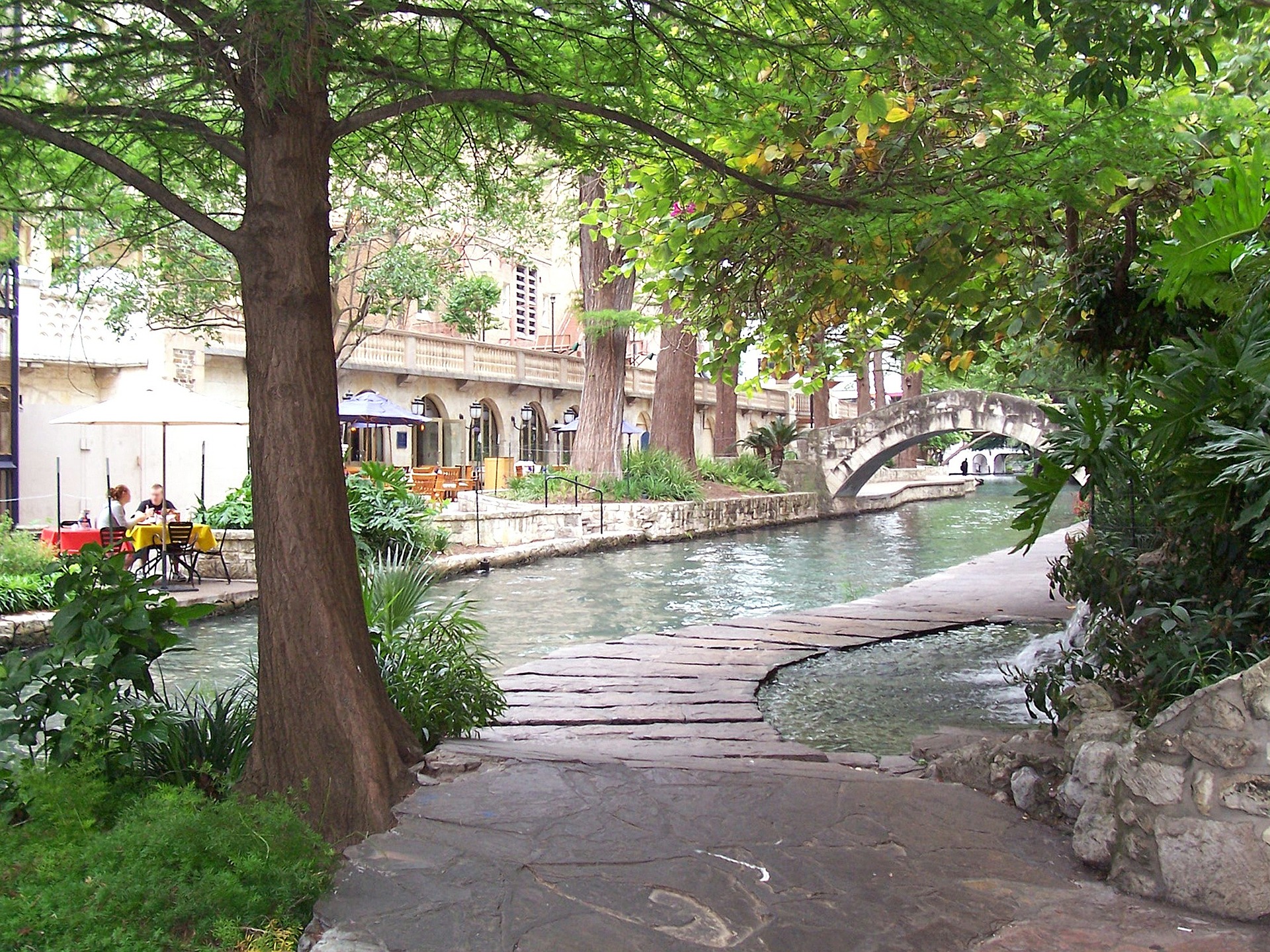 View of the San Antonio Riverwalk with a couple sitting riverside at an establishment.